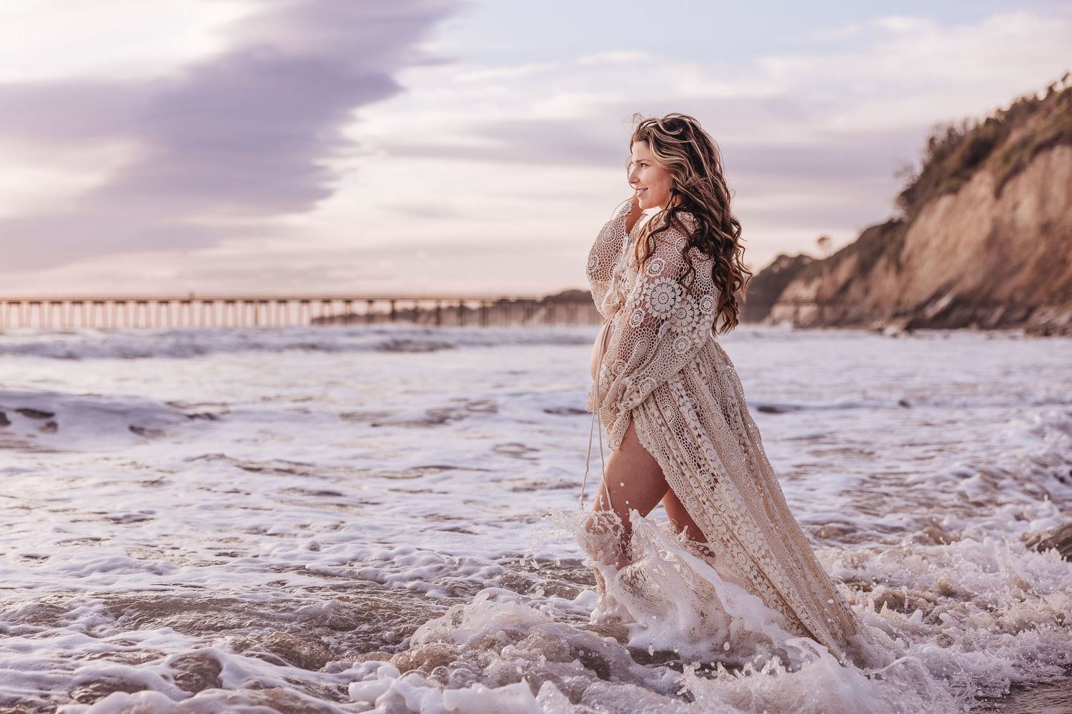 Flowing white lace dress splashes through ocean waves with dramatic cliff backdrop.