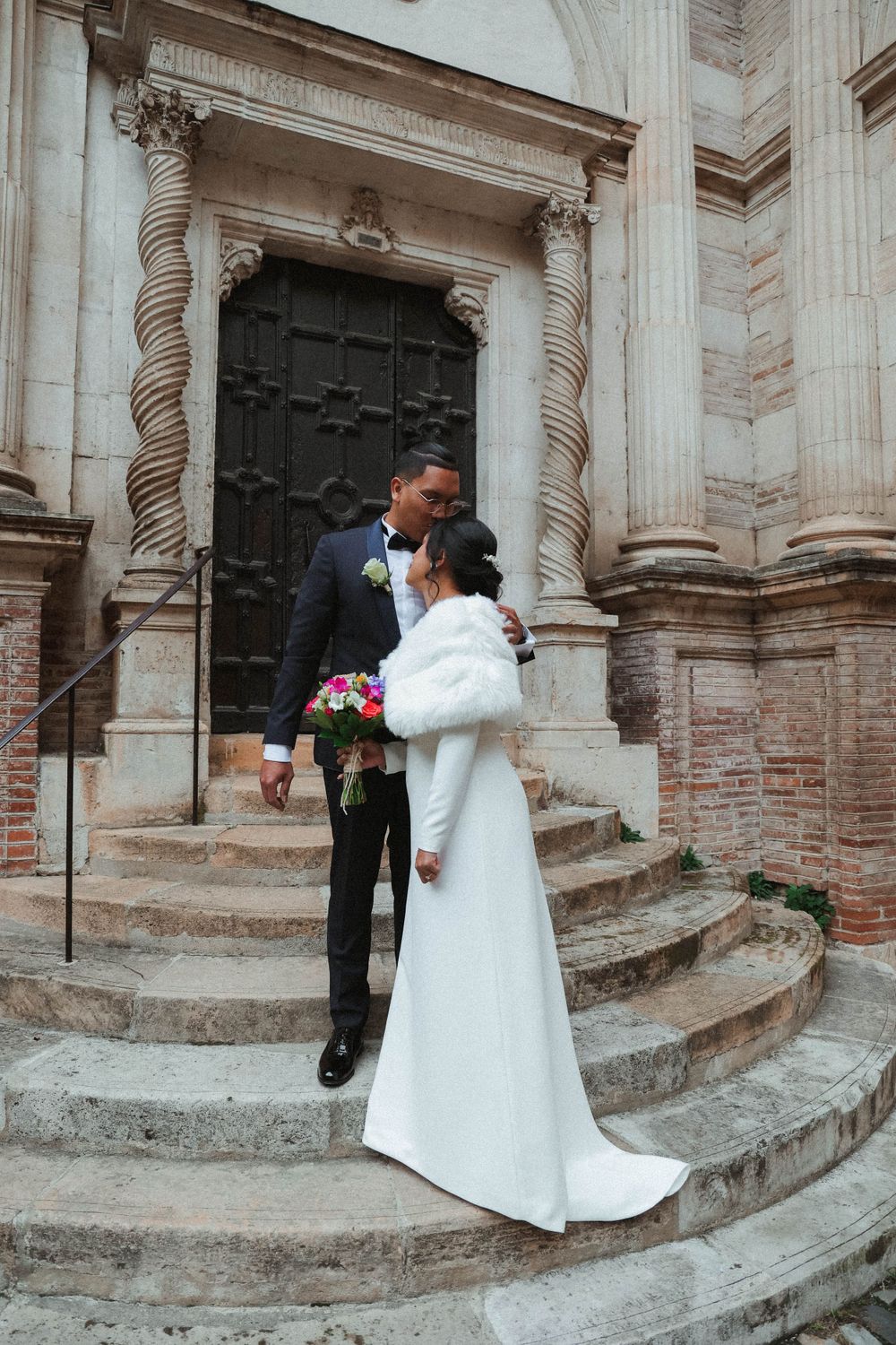 A couple in wedding attire stands on stone steps leading to an ornate church door.