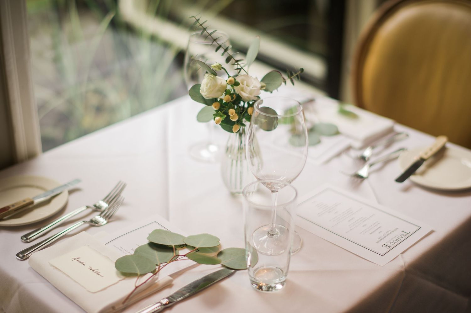 An intimate place setting with delicate floral arrangement and eucalyptus leaves on white tablecloth.