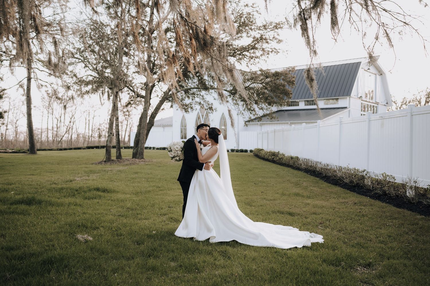 Wedding portrait under moss-draped trees with white fence backdrop.