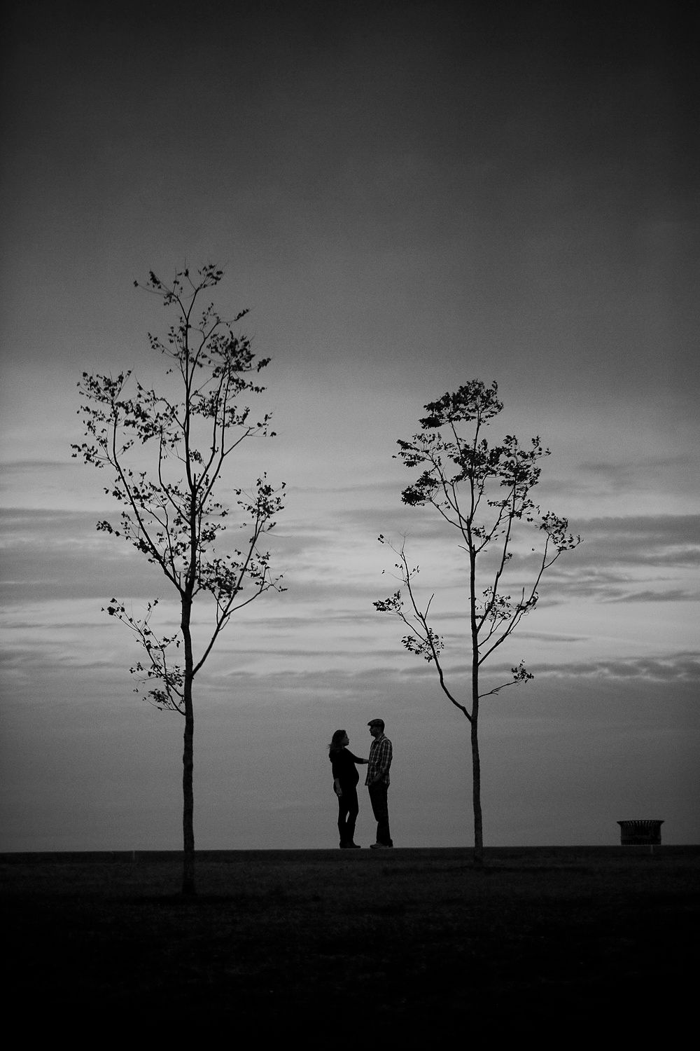 Silhouettes of two people standing between trees against dramatic sunset sky.