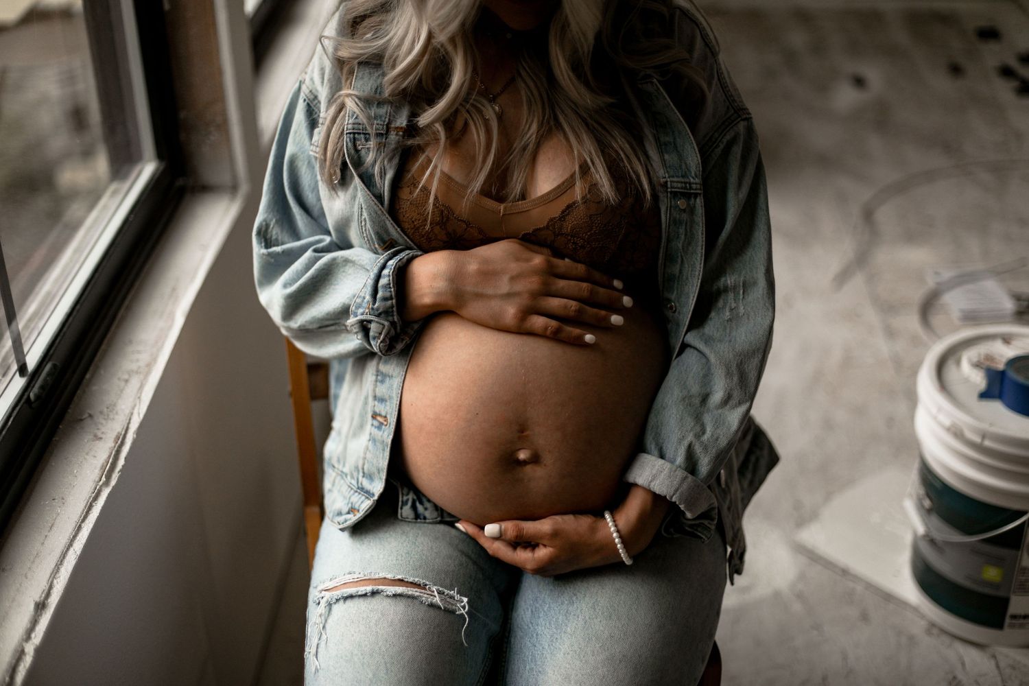 Intimate maternity moment by window with denim and natural lighting.