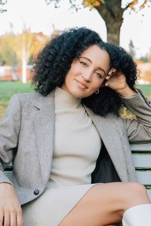 Young woman with curly hair in gray blazer sitting relaxed on park bench in autumn.