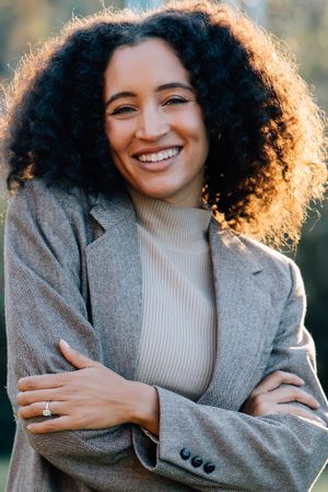 Smiling woman with curly hair and crossed arms wearing gray blazer outdoors in golden light.