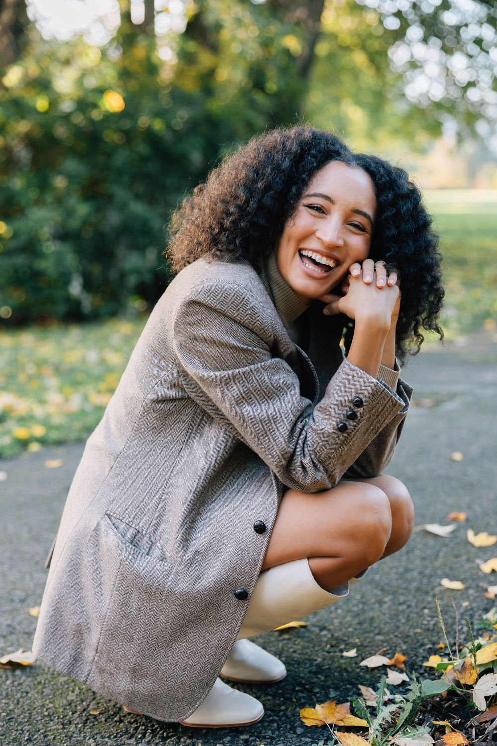 A joyful young woman with curly hair crouches outdoors in a beige blazer, laughing in an autumn park.