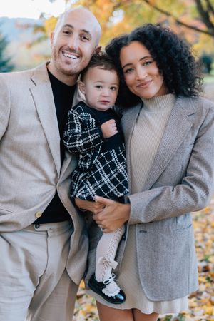 A smiling family of three poses together outdoors surrounded by colorful autumn leaves in a park.