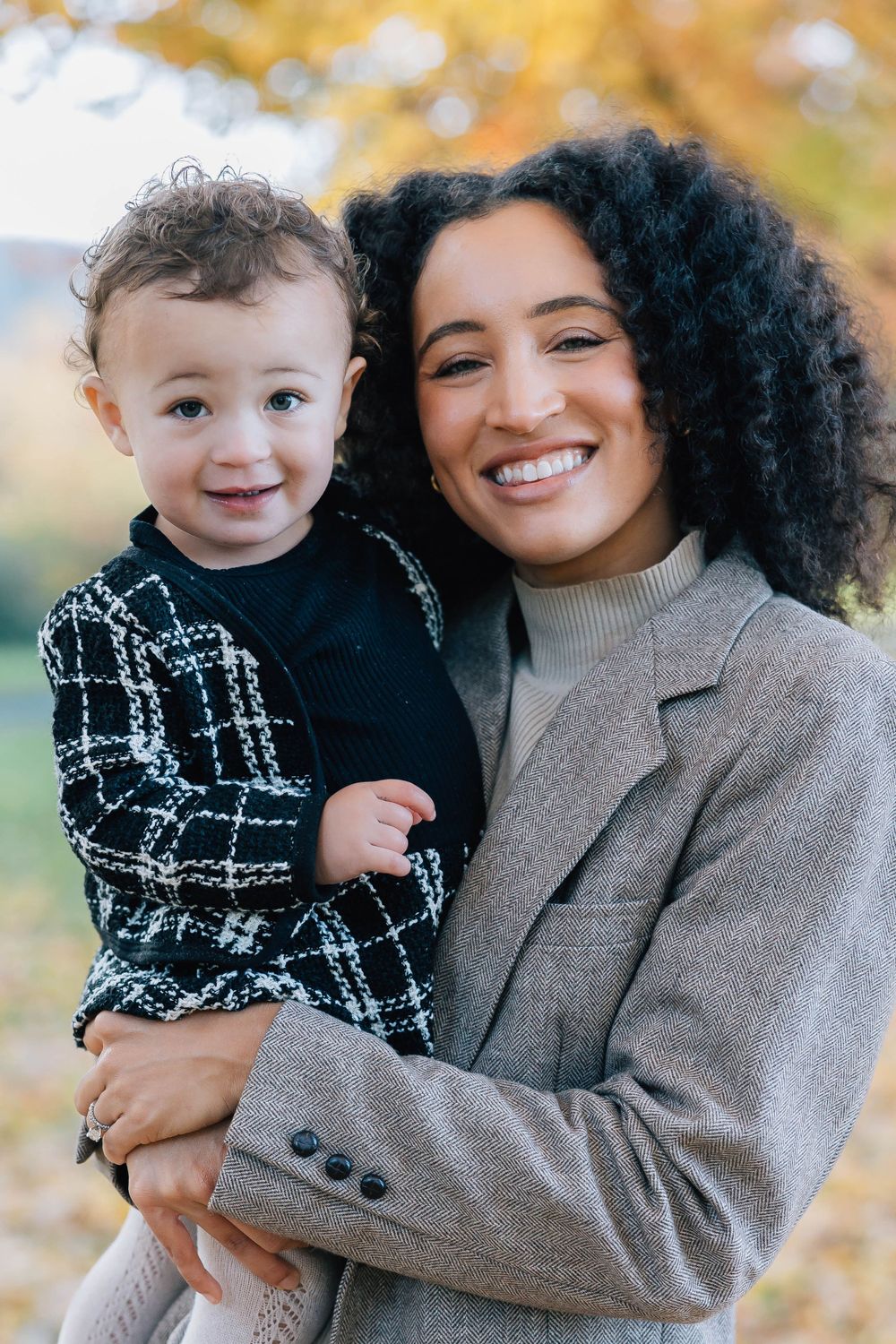A beaming mother with curly hair holds her adorable toddler close, surrounded by golden fall foliage.