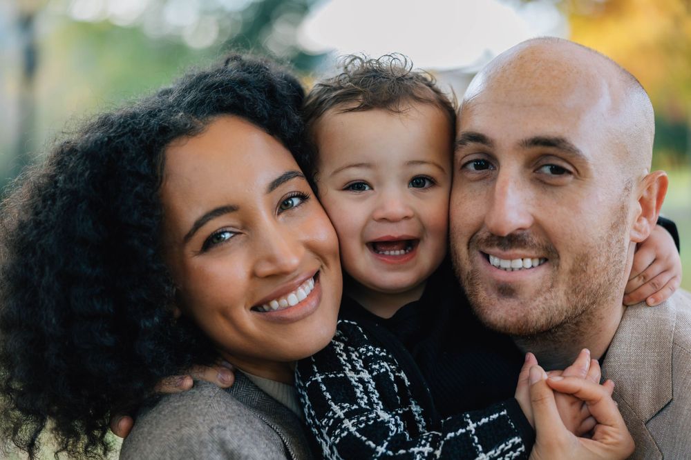 Happy mixed-race family portrait with smiling parents holding their laughing toddler outdoors in autumn.