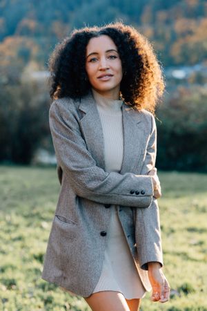 Confident young woman with curly hair in gray blazer posing in green outdoor field.
