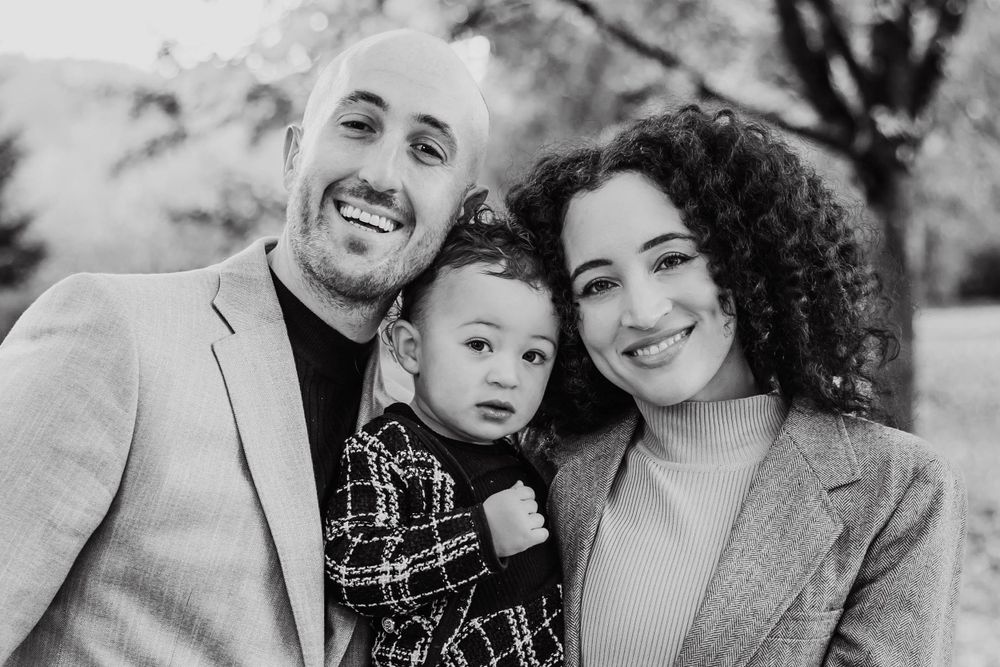 A black and white close-up portrait of a smiling couple with their young toddler in an outdoor setting.