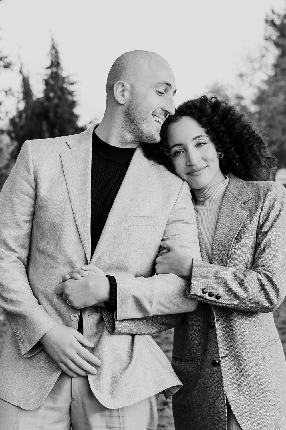 A black and white portrait of a loving couple embracing outdoors, the woman resting her head on the mans shoulder.