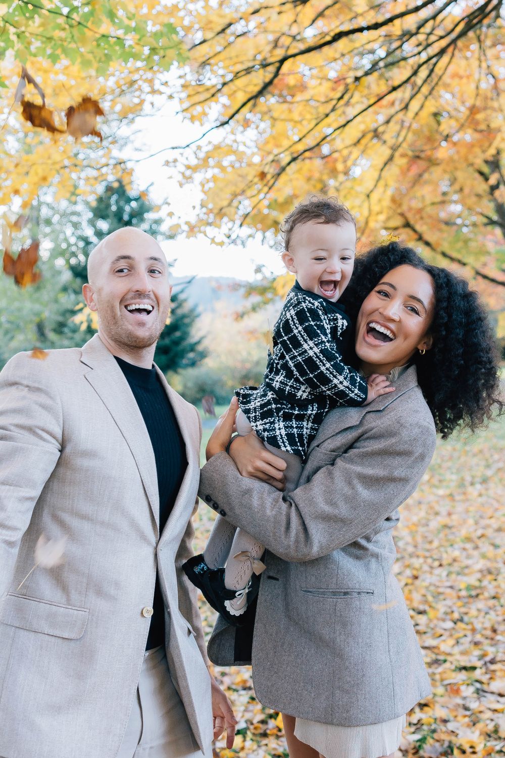 A laughing family of three enjoys a joyful moment together beneath golden autumn tree branches in a park.