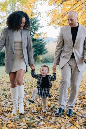 Happy interracial family walking hand-in-hand with toddler through colorful autumn park.