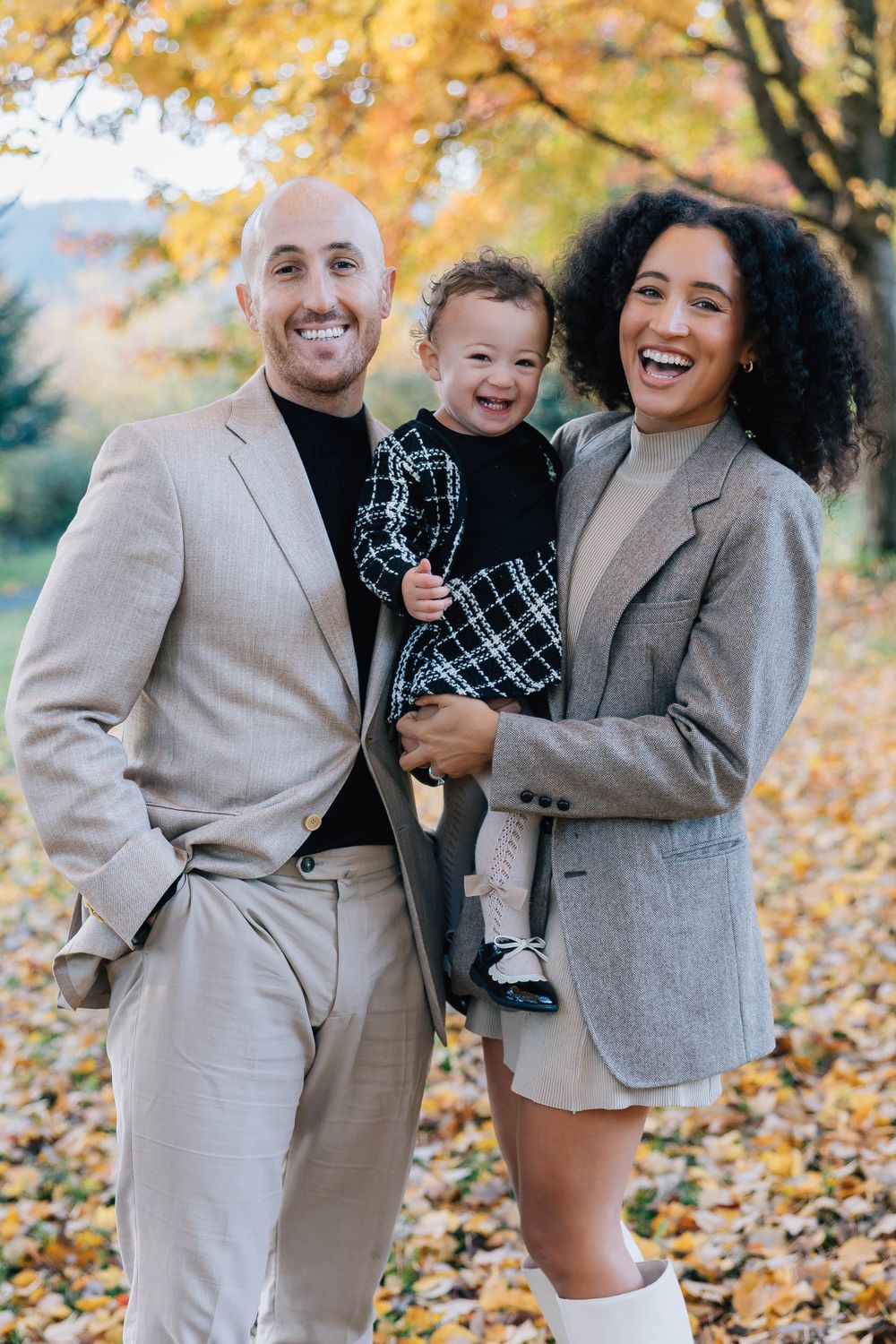 A joyful family of three laughs together among vibrant yellow fall foliage in an outdoor park setting.
