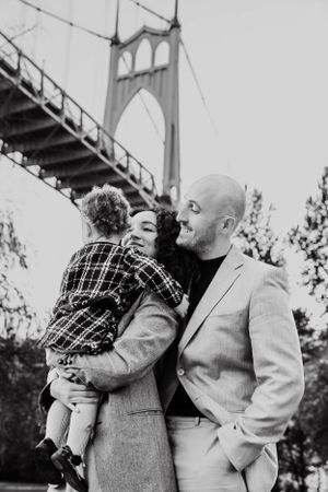 A black and white photo of a couple sharing a tender moment with their child beneath a large bridge.