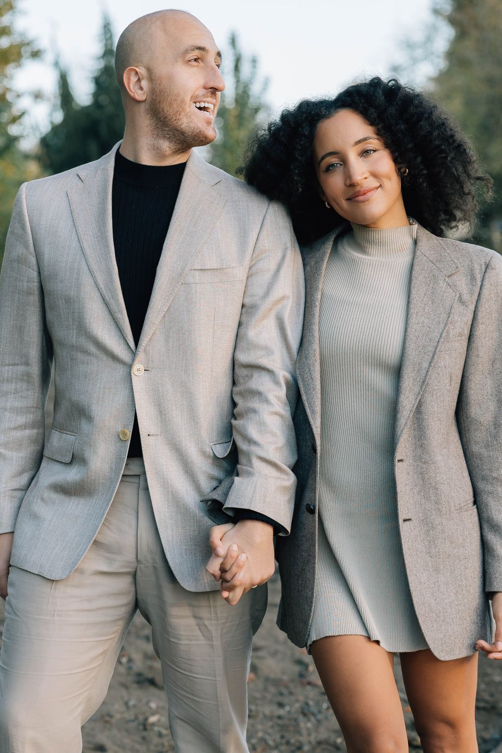 A stylish couple in matching beige blazers walks hand in hand through a park in autumn.