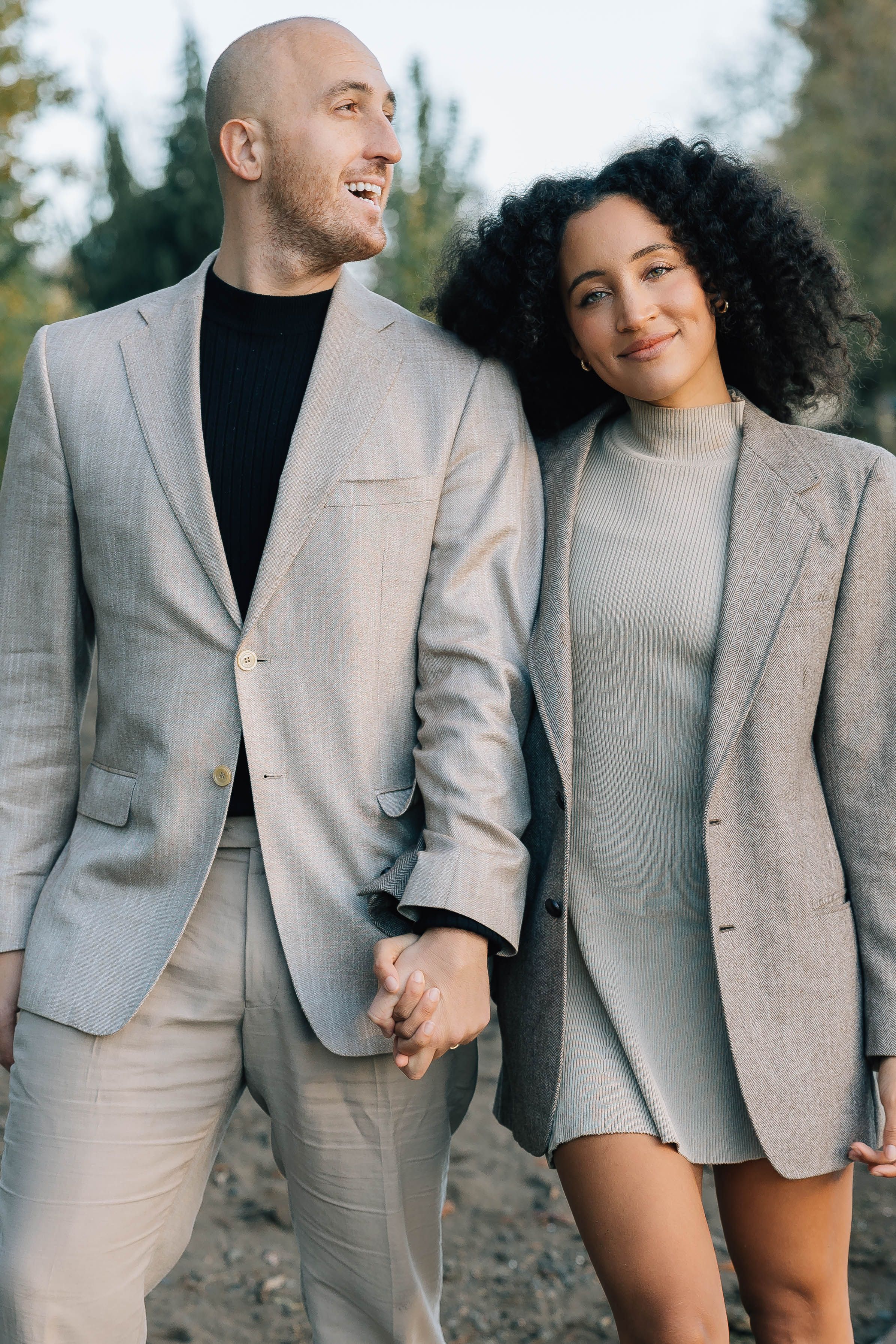 A stylish couple in matching beige blazers walks hand in hand through a park in autumn.