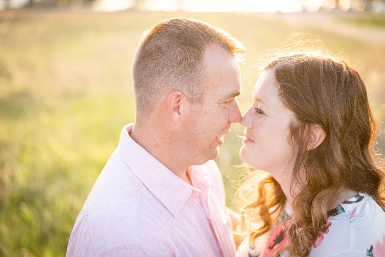 A romantic couple poses closely together in a sunlit field at sunset.
