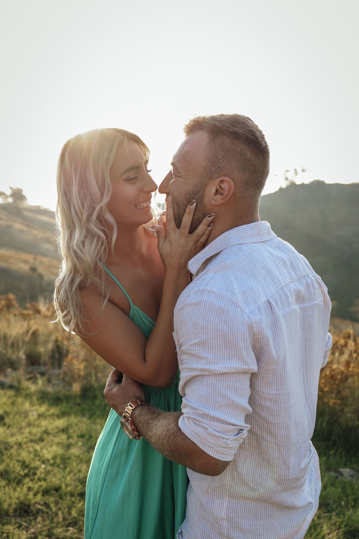 A couple sharing a romantic moment outdoors at sunset, woman in teal dress touching mans face in a sunlit field.