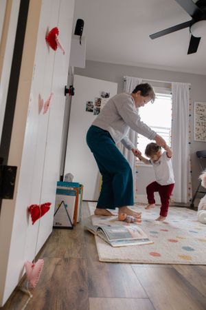 Sioux City, Iowa photographer. Parent helping toddler take first steps on a colorful rug in a decorated nursery room.