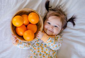 Sioux City, Iowa photographer. Smiling baby girl lying next to a wooden bowl of oranges wearing an orange print onesie.