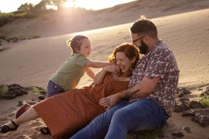 Sioux City, Iowa photographer. Happy family laughing together on sandy beach dunes at golden hour sunset in casual outdoor portrait.