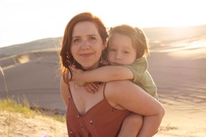 Sioux City, Iowa photographer. Mother carrying toddler on her back smiling at camera with sandy dunes glowing in sunset behind them.