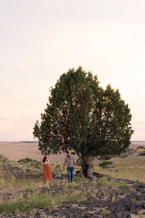 Sioux City, Iowa photographer. Family of three standing under large green tree in open landscape field during soft evening light.