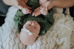 Sioux City, Iowa photographer. Newborn baby in green outfit and white bow headband lying on cream lace blanket with parents' hands.