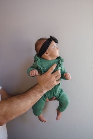 Sioux City, Iowa photographer. Father holding newborn baby dressed in green ruffle onesie and black bow headband against gray wall.