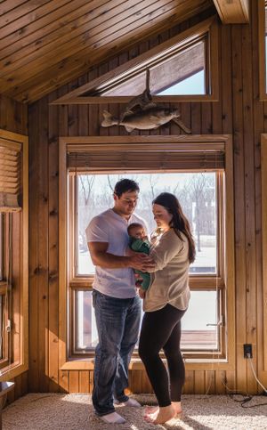 Sioux City, Iowa photographer. Young couple holding their newborn baby by a sunlit window in a cozy wood-paneled cabin.
