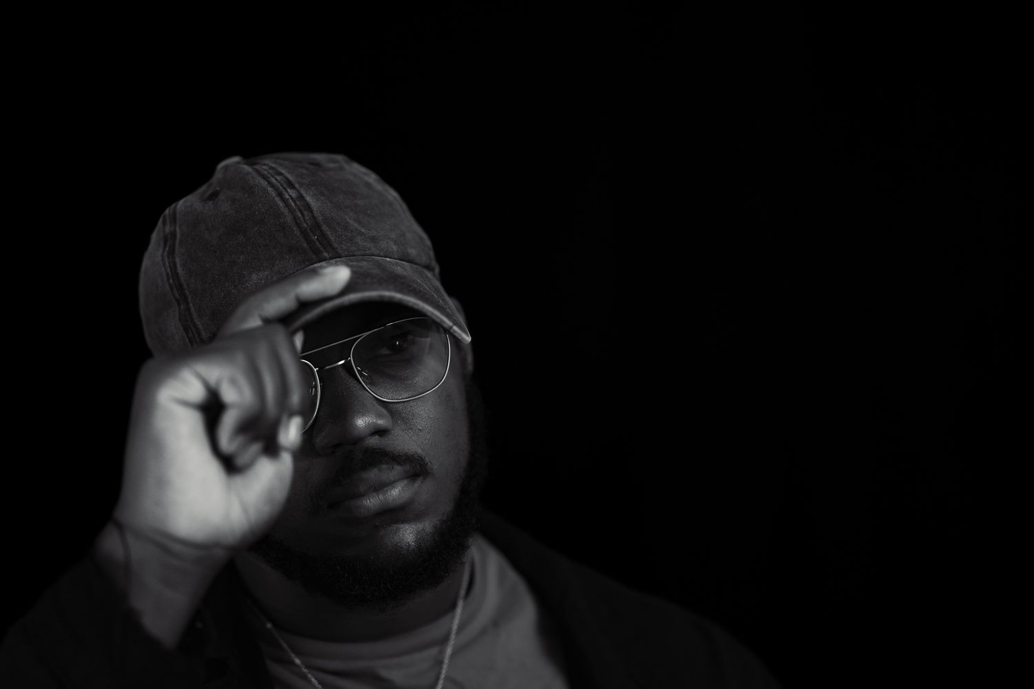 A moody black and white portrait shows a person adjusting their cap while wearing glasses.