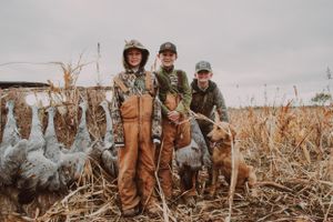 Hunting scene with figures in brown overalls posing with their dog in a field of dried corn.
