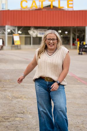 Person in cream sweater and jeans posing in front of red building with CATTLE sign.