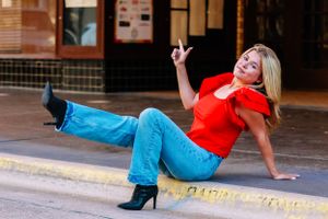 Playful fashion pose on curb wearing red top and blue jeans with black boots.
