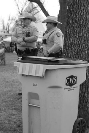 Two officers in cowboy hats stand beside a CWS waste container in black and white.
