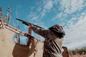 Hunter in camouflage taking aim with shotgun against blue sky.