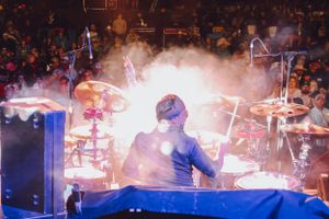 Concert drummer performing behind drum kit with dramatic lighting and smoke effects.