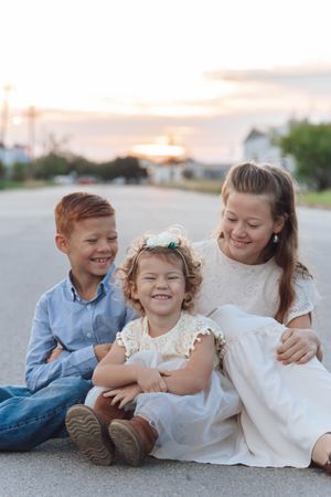 Three children sit together on a road at sunset sharing smiles and laughter.