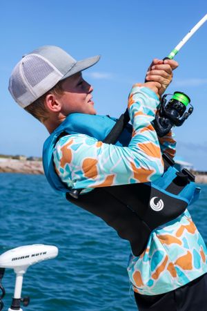 Angler in patterned UV protection gear reeling in catch on sunny fishing trip.