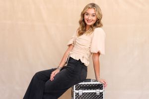 Stylish woman in cream top and black pants poses on metallic makeup case against beige backdrop.