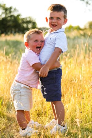 Two young brothers hugging and laughing in a sun-lit field of tall grass.