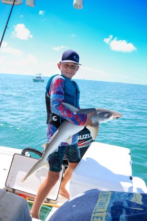 Young fisherman proudly displays caught shark on boat deck in bright blue waters.