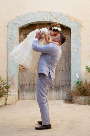 Wedding couple performs playful lifting poses against a weathered wall backdrop.