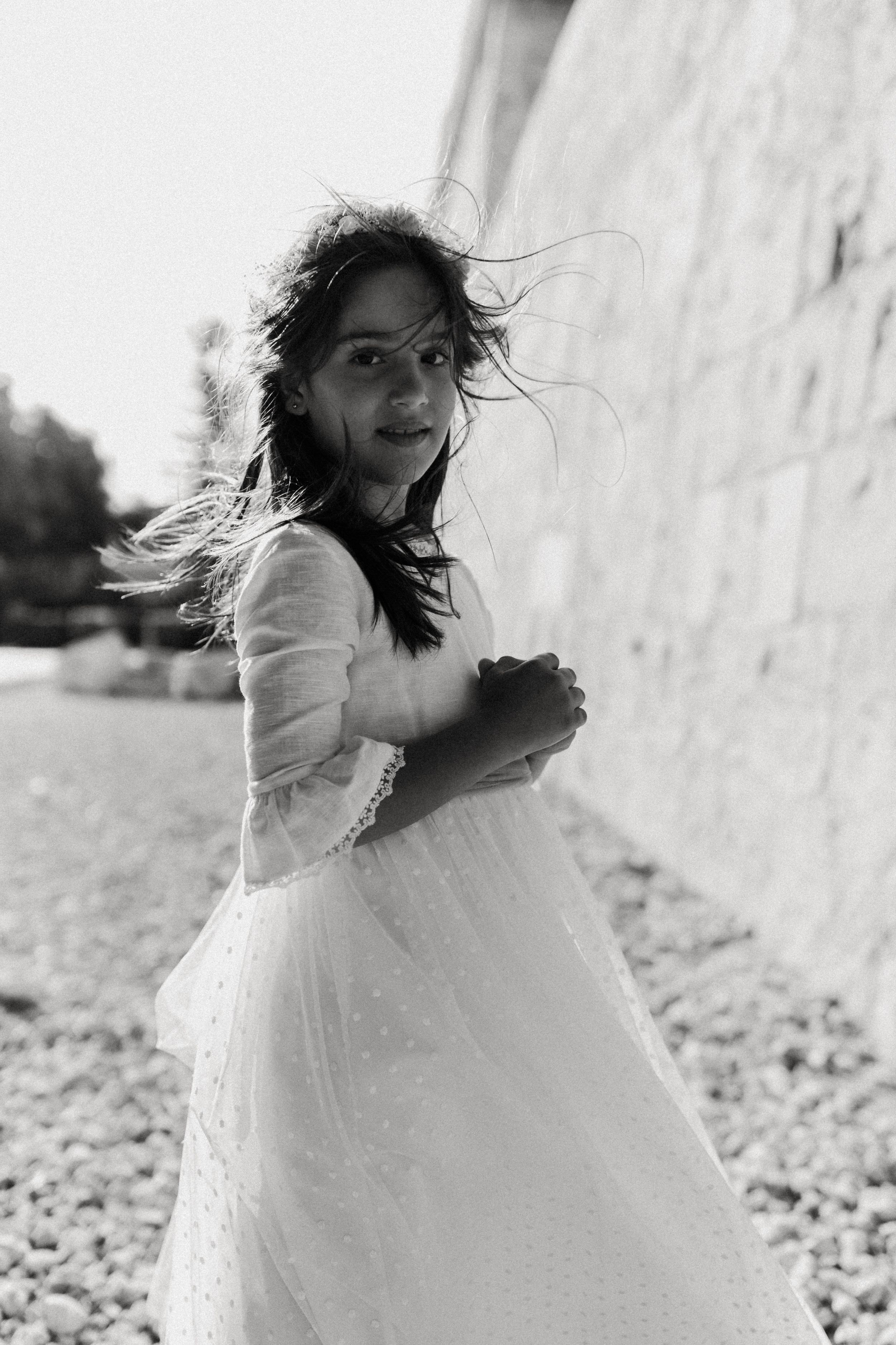 Young person in white dress twirls against stone wall backdrop.