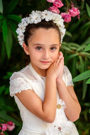 A child in white lace dress and floral crown poses against green foliage.
