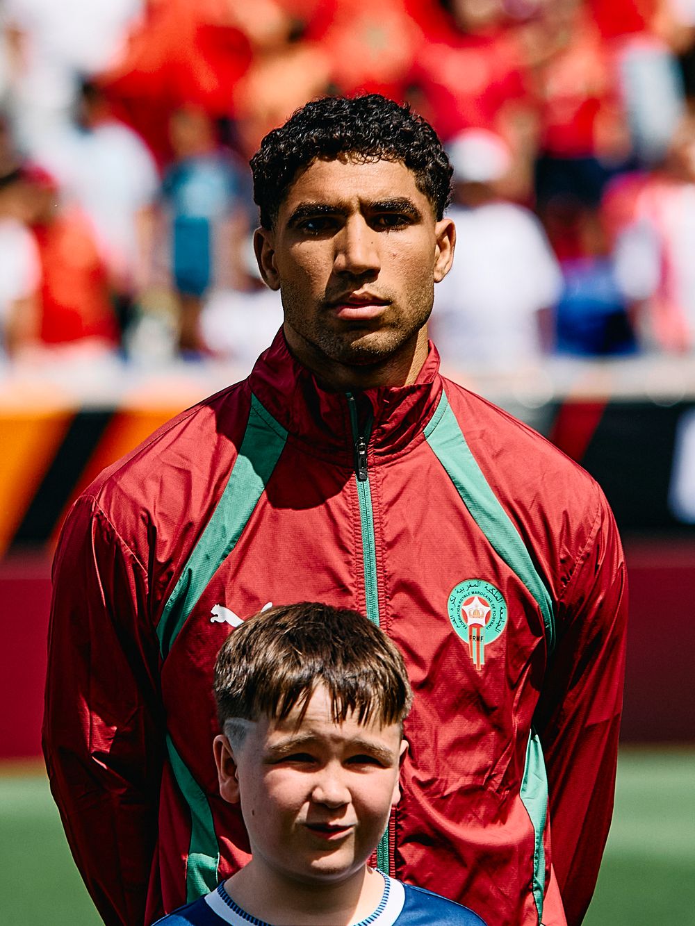 A Moroccan soccer player in a red and green Puma jacket stands behind a young child mascot before a match.