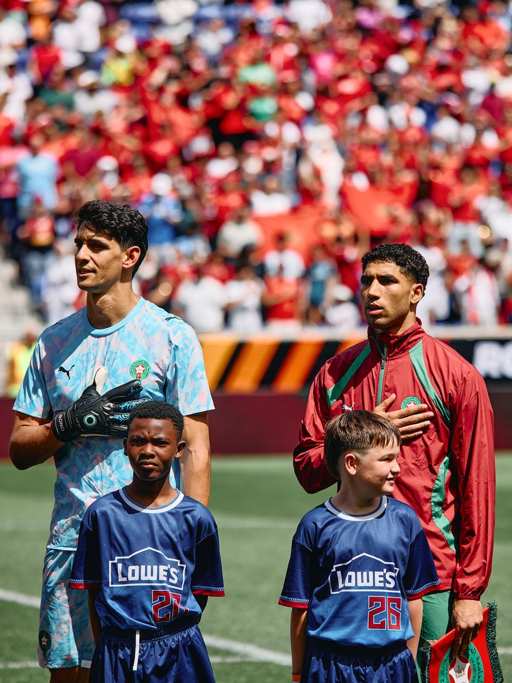 Two Morocco soccer players stand with young mascots during the national anthem before an international match at a packed stadium.