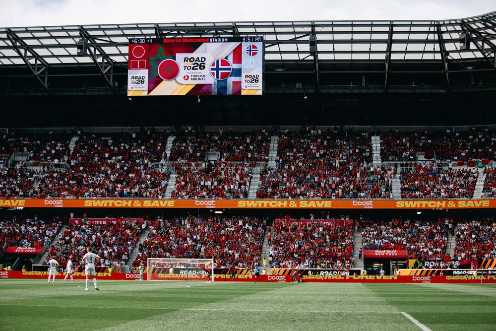 Soccer stadium filled with fans during a match, featuring a large scoreboard displaying the Road 26 logo under sunny skies.