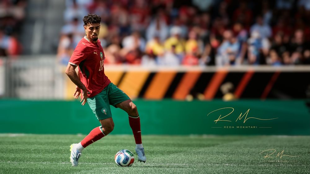 A soccer player in a red and green Portugal kit dribbles a ball on the field during a sunny outdoor match.
